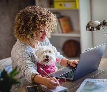 Woman taking notes while looking at her laptop and holding her dog