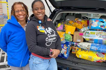 Two women standing in front of their car trunk full of pet food donations