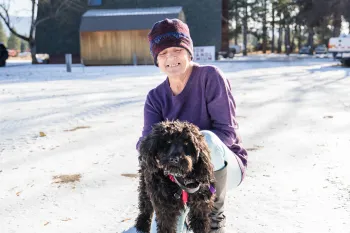 A woman in a purple beanie and shirt holds a black, curly haired dog in the snow