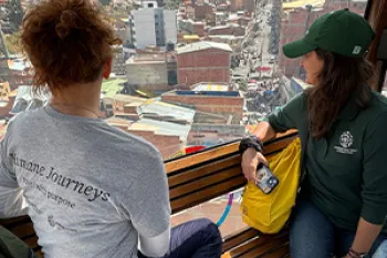 Two people look out a window over a dense population center with mountains in the background.