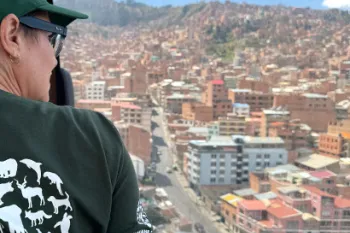 A person wearing glasses, a green hat and a green shirt look out a window over a dense city.